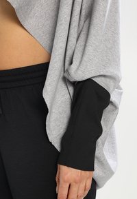 Gros plan sur une personne portant un haut à manches longues côtelées noir sous un top gris ample, et un pantalon à taille élastique noir montrant une partie de la taille.