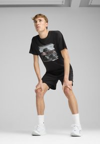 Sort t-shirt med korte ærmer, der har et grafisk print, parret med sorte shorts og hvide sneakers. Modellen poserer med hænderne på knæene.