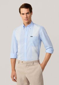 Chemise bleu clair à manches longues avec des panneaux �à rayures horizontales et une poche poitrine ; les manches pleines sont retroussées. Portée avec un pantalon beige.