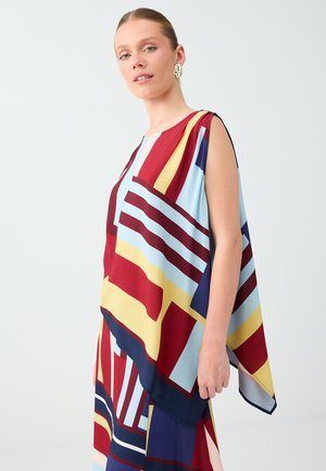 Femme portant un haut sans manches coloré avec des motifs géométriques en rouge, jaune, bleu et bleu clair, debout devant un fond uni.