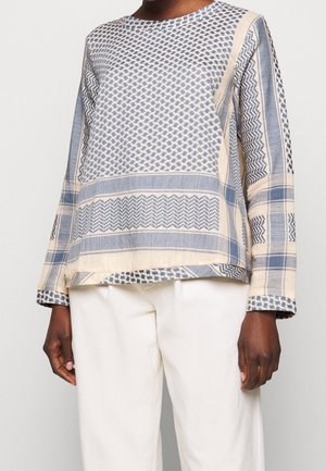 Person iført en langærmet beige top med blå geometriske mønstre og off-white bukser, der står mod en ensfarvet baggrund.