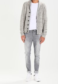 Cardigan grigio lavorato a maglia con colletto a scialle, chiusura a bottoni, abbinato a jeans chiari strappati e sneakers bianche.