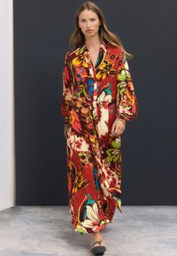 Vestito lungo a fiori nei vibranti colori rosso, giallo e verde. Presenta una chiusura con bottoni, scollo con colletto e maniche ampie per una vestibilità comoda.