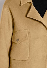 Kamel-wollmantel mit breitem Reverskragen und Klappentasche, die auf der linken Seite mit einem Metall-Druckknopf gesichert ist.