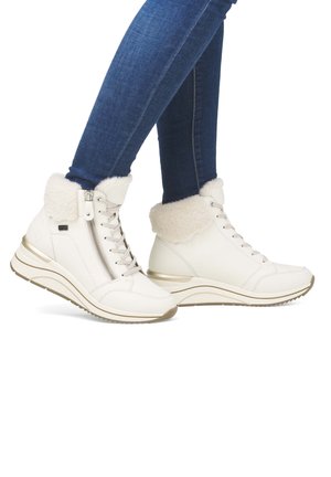 Legs in dark blue jeans wearing white high-top sneakers with fur lining, lace-up front, side zipper, and thick sole on a white background.