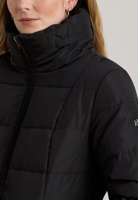 Svart vadderad puffjacka med hög krage, utrustad med quiltade sektioner och dragkedjestängning. Vänsterärm visar logotypen i kontrasterande text.