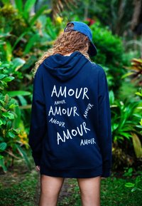 Sötétkék kapucnis pulóver puha anyagból, nagy kenguruzsebbel. A hátsó részén a "AMOUR" felirat különböző fehér betűtípusokkal van szétszórva.