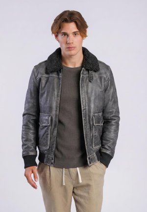 Młody mężczyzna o jasnobrązowych włosach, ubrany w czarną skórzaną kurtkę z kołnierzem z owczej wełny, ciemny sweter i beżowe spodnie, stoi na jednolitym tle.