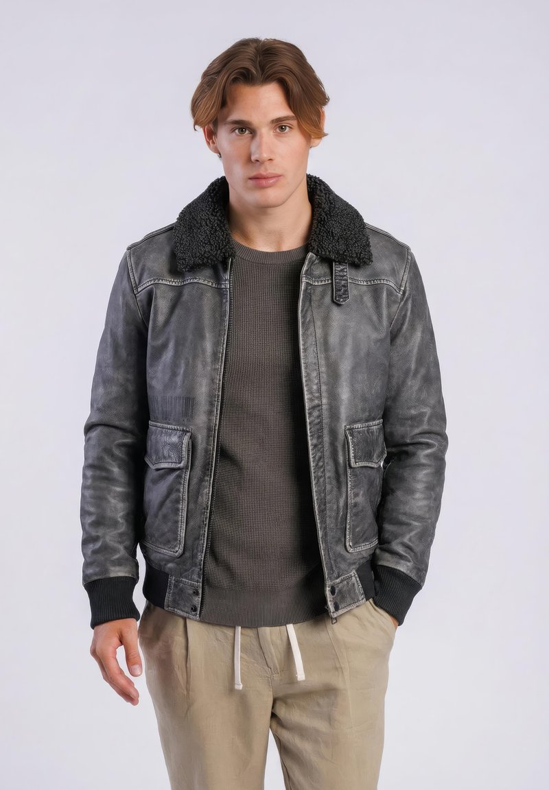 Jonge man met lichtbruin haar, gekleed in een zwart lederen jack met een kraag van schapenvacht, een donker trui en beige broek, staand tegen een effen achtergrond.