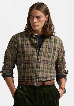 Camicia a maniche lunghe a quadri nei colori verde, marrone e blu navy, con logo sul petto. Indossata sopra un maglione scuro e abbinata a pantaloni di velluto a coste scuri.