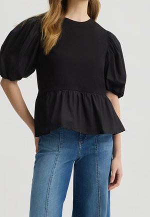 Kvinde iført sort peplum-top med pufærmer kombineret med blå højttaljede jeans, stående mod en ensartet lys baggrund.