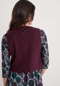 Burgundski vuneni prsluk s jednostavnom stražnjom površinom, uparen s haljinom cvjetnog uzorka koja sadrži različite nijanse zelene i ljubičaste boje.