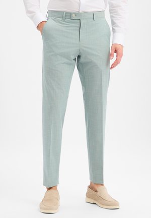 Man draagt lichtgroene op maat gemaakte broek met voorzakken, gecombineerd met beige instapschoenen en een wit overhemd.