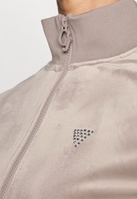 Veste beige à col haut zippée avec col côtelé et petit motif triangulaire de clous brillants sur la poitrine.
