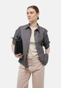 Chemise à rayures marron foncé et bleu clair, assortie à un haut beige et un pantalon rose clair, tenant un sac noir.