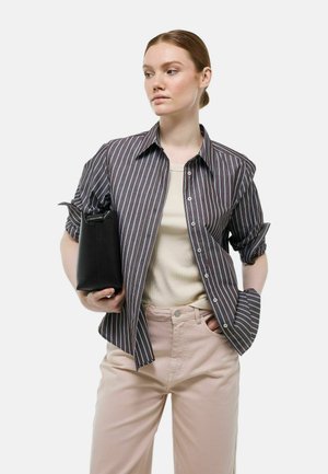 Chemise à rayures marron foncé et bleu clair, assortie à un haut beige et un pantalon rose clair, tenant un sac noir.