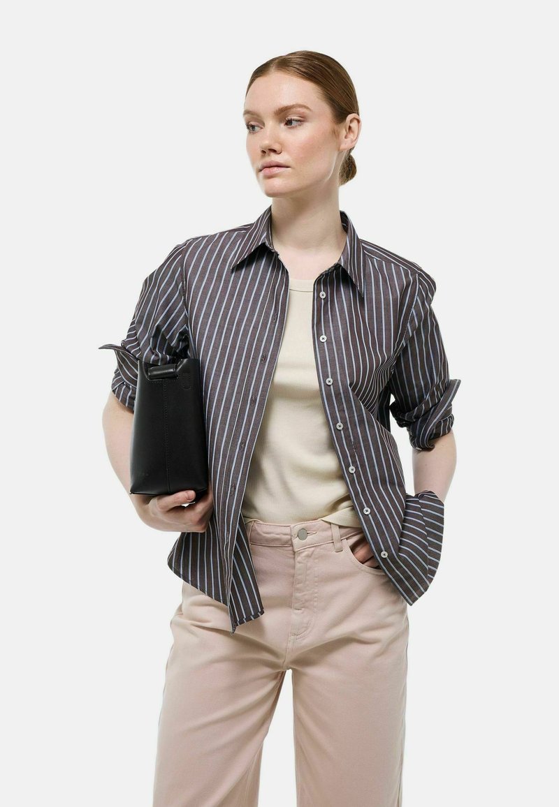 Chemise à rayures marron foncé et bleu clair, assortie à un haut beige et un pantalon rose clair, tenant un sac noir.