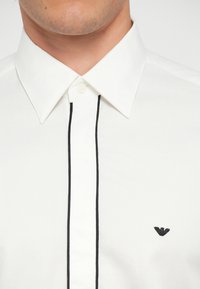 Biała koszula z kołnierzykiem z czarnymi przeszyciami wzdłuż zapięcia. Posiada małe czarne logo po lewej stronie klatki piersiowej.