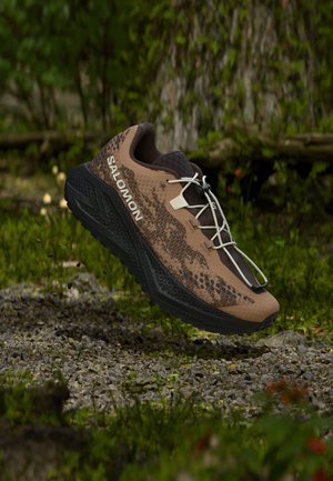 Καφέ και μαύρο παπούτσι trail τρεξίματος Salomon στον αέρα πάνω από χαλίκι και πράσινο βρύο έδαφος με φόντο το δάσος.