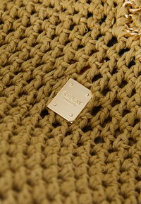 Tissu tissé de couleur moutarde avec un motif texturé et noué, comportant une étiquette en métal doré embossée avec "REPLAY" et "SINCE 1981."
