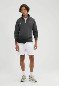 Szara bluza na zamek z logo, w połączeniu z białymi szortami. Czarna torba na biodra, a na nogach neutralne sneakersy z zielonymi akcentami.