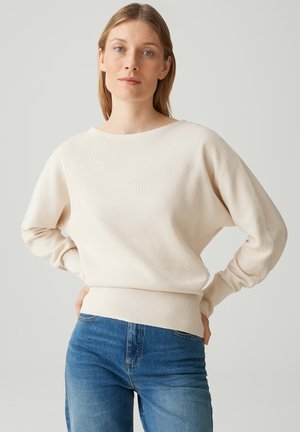 Donna con pelle chiara e capelli lisci, che indossa un maglione crema a maniche lunghe e jeans blu, in piedi con le mani sui fianchi contro uno sfondo semplice.