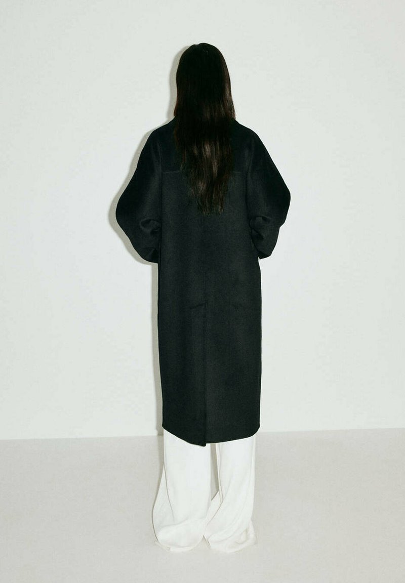 Personne aux longs cheveux foncés portant un long manteau noir et un pantalon blanc à jambes larges, se tenant face à un mur uni gris clair.