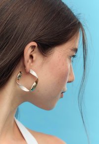 Guldfarvede og sølvfarvede hoops øreringe med et bølgemønster, der har en blank overflade og en reflekterende finish, båret af en model mod en blå baggrund.