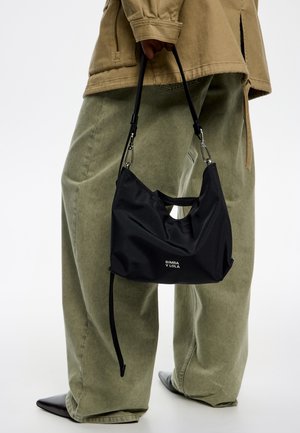 Bolso de nylon negro con un diseño curvado, que cuenta con una correa para el hombro y un logo. Se lleva con pantalones holgados de color verde oliva y zapatos de punta.