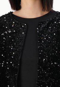 Giacca nera con paillettes e una morbida texture di velluto, caratterizzata da un colletto rotondo e un design aperto sul davanti, indossata sopra un top nero.