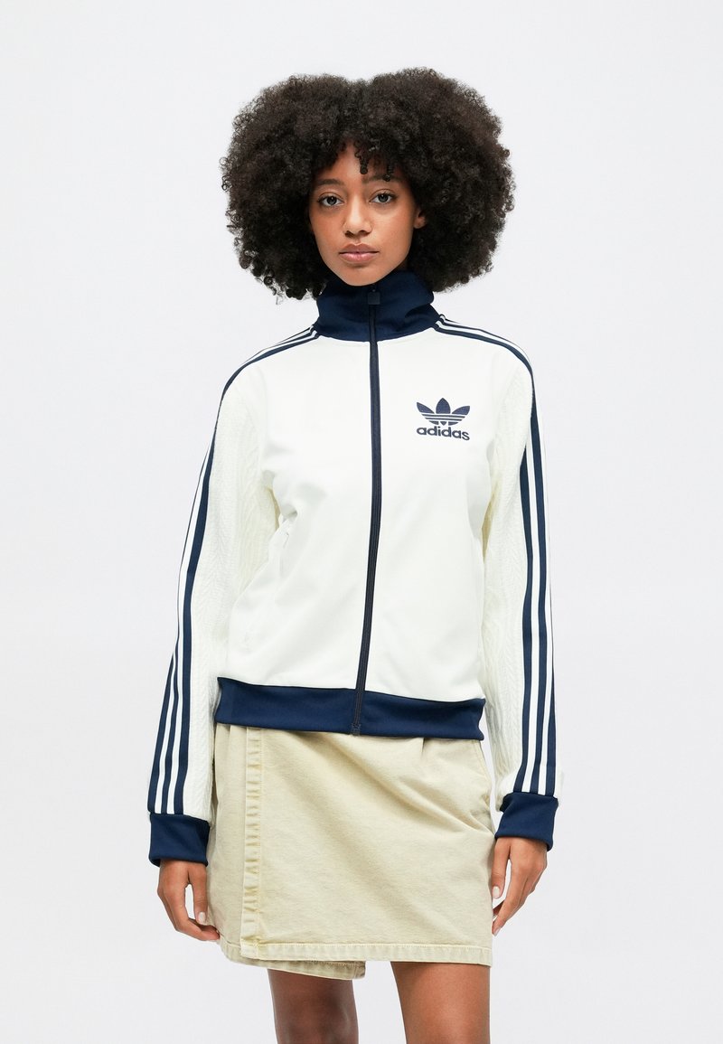 White jacket with navy accents, high collar, full zip, and three-stripe design on sleeves. Soft texture with a fitted silhouette and logo detail.