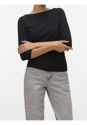 Personne portant un haut noir à manches trois-quarts et un jean gris clair taille haute, les bras croisés sur le torse devant un fond uni.