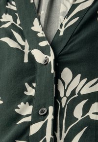 Indumento verde a motivo floreale con design a scollo a V, tessuto testurizzato e bottoni neri. Il motivo è composto da grandi foglie bianche e fiori.