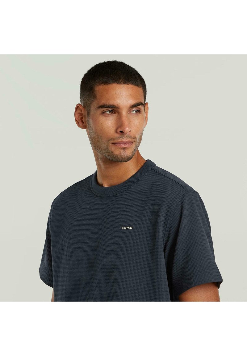 Hombre con cabello corto vistiendo una camiseta de manga corta texturizada G-Star azul oscuro, mirando ligeramente hacia su derecha con un fondo liso.