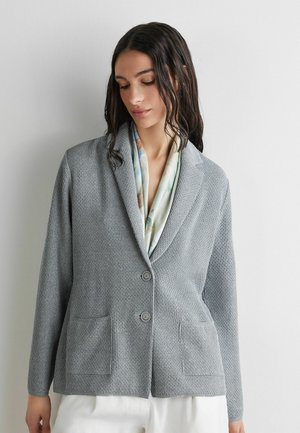 Donna che indossa un blazer grigio chiaro con texture sopra una sciarpa fantasia e pantaloni bianchi, in piedi davanti a uno sfondo bianco semplice.