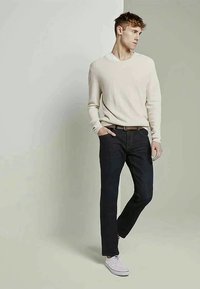 Conjunto masculino com um suéter de malha creme e jeans escuros. Ténis brancos completam o visual. O suéter tem um design com decote em V.