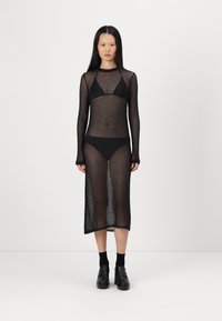 Černé pletené šaty s dlouhými rukávy a maxi délkou, pod nimiž se nachází černé bikini. Vyznačují se průsvitnou texturou a obtaženým střihem.