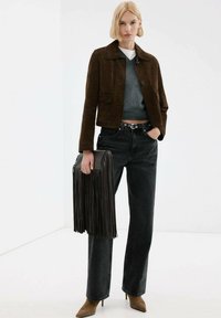 Veste en suède marron, pull gris, jean noir taille haute et bottines marron. Tenant un sac à main noir à franges. Fond simple.