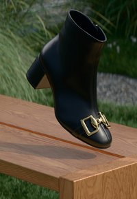 Botine negre din piele, cu vârf ascuțit, accent cu cataramă aurie și toc bloc de înălțime medie, așezate pe o suprafață din lemn.