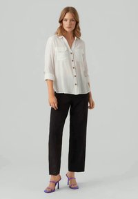 Camisa branca de botões com dois bolsos no peito, calças pretas ajustadas e saltos roxos com tiras, contra um fundo neutro.
