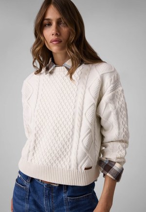 Femme portant un pull en maille blanche texturée par-dessus une chemise à carreaux avec les manches retroussées et un jean bleu sur un fond gris uni.