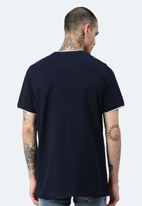 T-shirt in cotone blu navy con collo rotondo, maniche corte e bordi bianchi a contrasto sui lati delle maniche. Presenta tatuaggi visibili sulle braccia.
