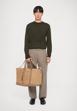 Bolso tote de ante marrón con dos asas y un bolsillo con cremallera, combinado con un suéter verde oscuro y pantalones beige con estampado. Se llevan zapatos negros.