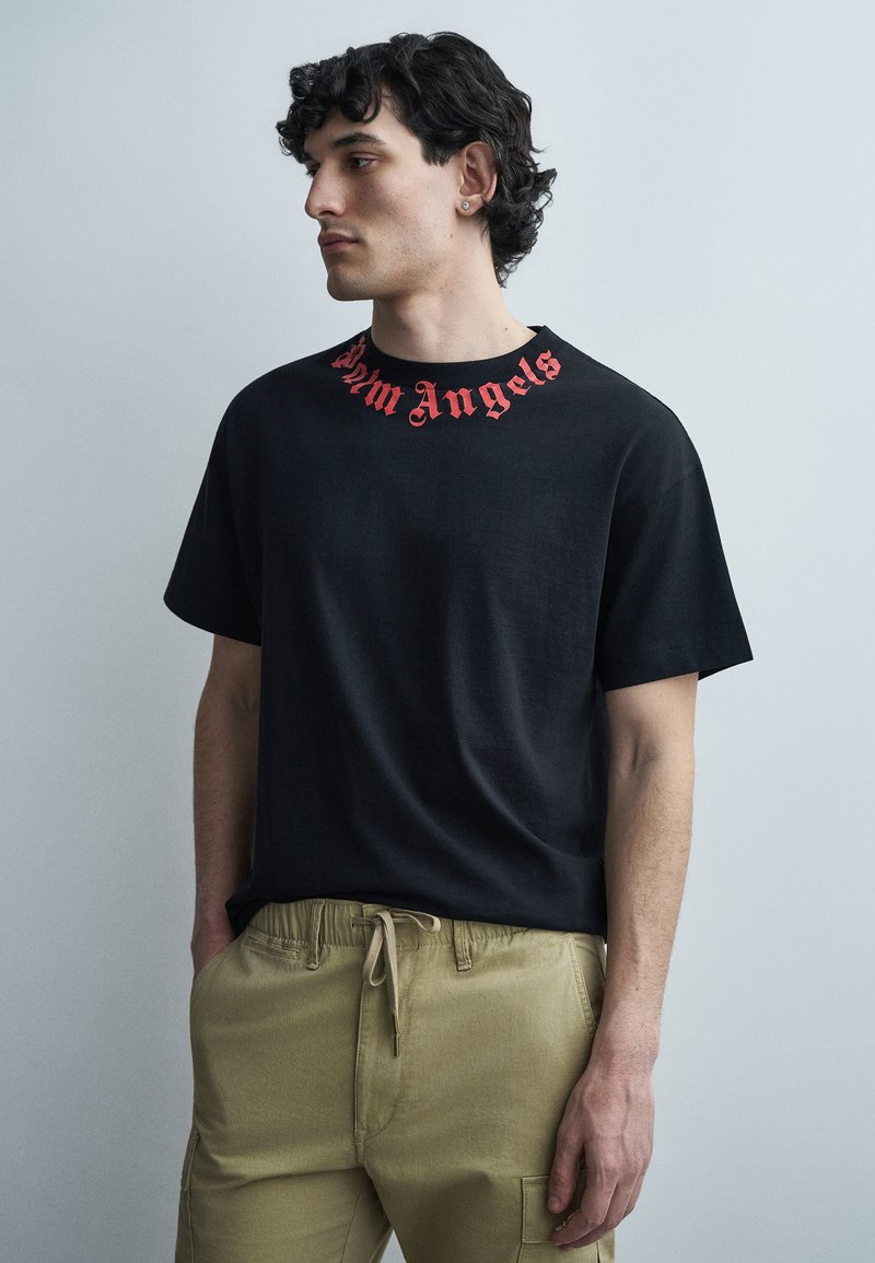 Fiatal férfi göndör sötét hajjal, fekete pólót visel, amelynek nyakánál piros "Palm Angels" felirat található, és bézs színű, zsinórral állítható nadrágot, kezét zsebében tartja.