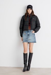 Svart puffjakke, denim mini-skjørt med knappdetaljer og knelange svarte støvler. Modell med en matchende svart caps.