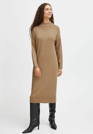 Braunes Strickkleid mit hohem Hals, langen Ärmeln und geradem Schnitt, getragen mit eng anliegenden, schwarzen Stiefeln bis zum Knie. Glatte Textur.