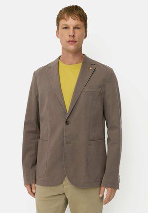 Mand iført en taupe blazer over en gul skjorte og beige bukser, stående mod en hvid baggrund, vendt fremad.