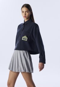 Gekürzte marineblaue Denimbluse mit einer aufgesetzten Tasche und grünen gestickten Details, kombiniert mit einem hellgrauen, plissierten Rock.
