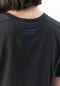 Pessoa com cabelo escuro curto, vestindo uma t-shirt preta com o texto roxo "AMERICAN VINTAGE" nas costas, abaixo da gola.