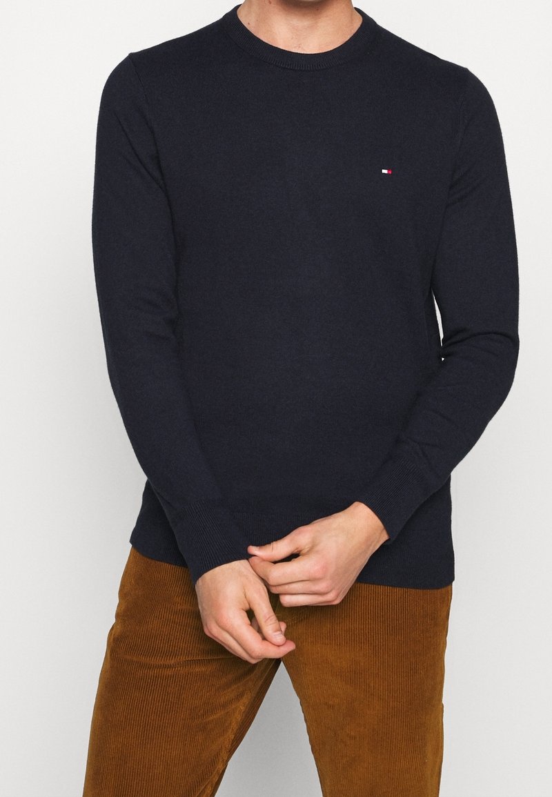 Maglione lavorato a maglia blu navy con colletto a girocollo, caratterizzato da maniche lunghe e un piccolo logo sul petto, abbinato a pantaloni in velluto a coste.